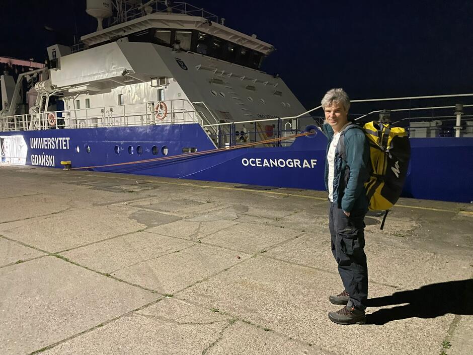 Na zdjęciu widoczny jest mężczyzna z szarymi włosami, ubrany w ciemne spodnie i zieloną kurtkę, stojący na nabrzeżu obok statku OCEANOGRAF  należącego do Uniwersytetu Gdańskiego. Mężczyzna ma na plecach duży żółto-czarny plecak i patrzy w stronę aparatu z uśmiechem.