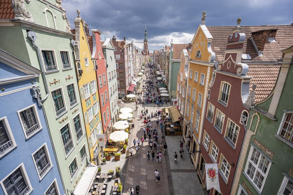 Na zdjęciu przedstawiona jest ulica w Gdańsku, widziana z góry. Po obu stronach ulicy znajdują się charakterystyczne, kolorowe kamienice o zdobionych fasadach. Kamienice są pomalowane na różne kolory: niebieski, zielony, żółty, czerwony i pomarańczowy. Ulica jest wypełniona spacerującymi ludźmi, a wzdłuż niej ustawione są białe parasole przy kawiarnianych stolikach i straganach. W tle, na końcu ulicy, widać wieżę kościoła z zegarem. Nad miastem zbierają się ciemne chmury, co sugeruje nadchodzącą burzę lub deszcz. Cała scena tętni życiem i jest pełna barw.