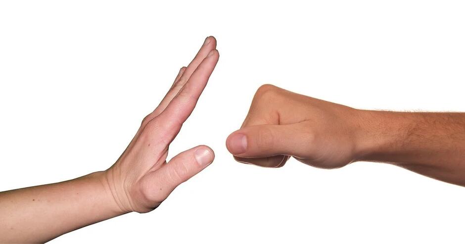 Na zdjęciu widzimy dwie dłonie na białym tle. Jedna dłoń jest zaciśnięta w pięść, a druga otwarta, wyciągnięta w geście powstrzymującym, symbolizującym sprzeciw wobec przemocy
