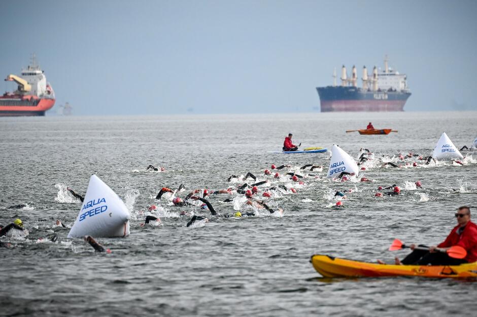 Na zdjęciu widać grupę pływaków uczestniczących w zawodach na otwartym morzu, nad którymi czuwają osoby na pontonie i kajakach. W tle można dostrzec kilka dużych statków. 