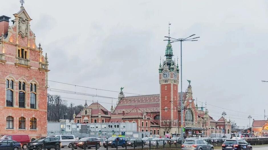 Na zdjęciu widać dworzec kolejowy Gdańsk Główny, charakterystyczny ze względu na swoją architekturę z wysoką wieżą zegarową. Przed dworcem znajduje się ruchliwa ulica z samochodami, a w tle można dostrzec tory tramwajowe.