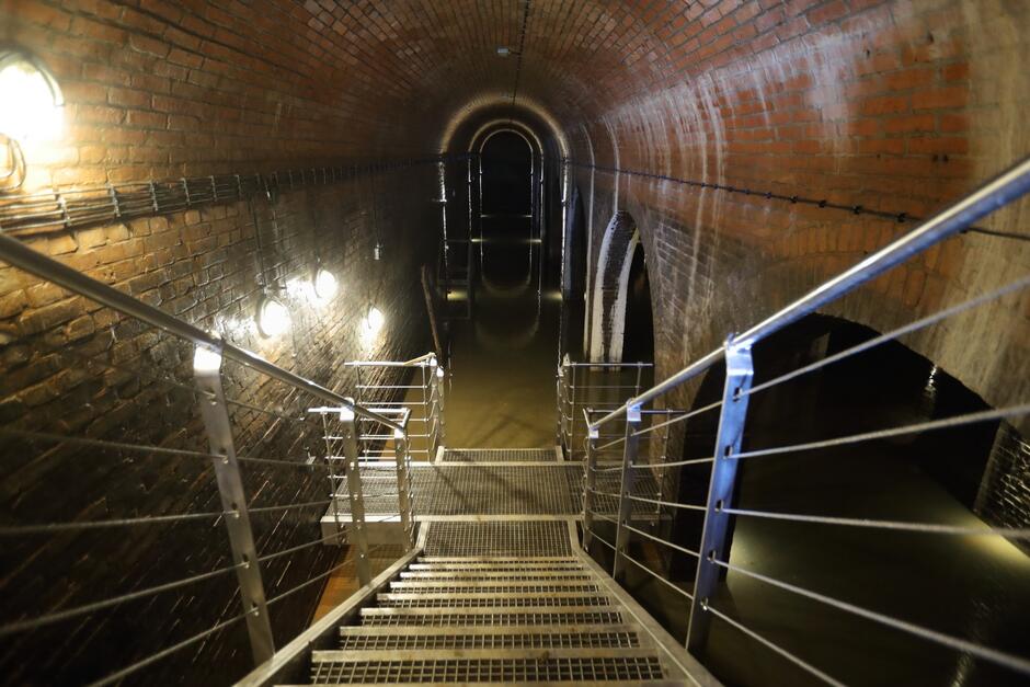 Stalowe ażurowe schody prowadzą w dół podziemnego zbiornika wodnego, którego łukowate ściany wykonane są z cegły. na posadzce lustro wody.