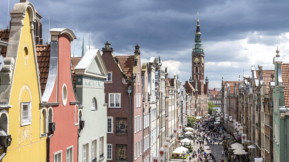 Na zdjęciu widzimy główną ulicę Gdańska, prowadzącą w kierunku wieży Ratusza Głównego Miasta, która dominuje w tle. Wieża ratusza jest ozdobiona zegarem i zieloną iglicą, a jej brązowa fasada kontrastuje z jasnym niebem. Nad miastem zbierają się ciemne chmury, co dodaje dramatyzmu scenerii. Po obu stronach ulicy znajdują się kolorowe kamienice o charakterystycznych, ozdobnych fasadach. Kamienice mają różne kolory: żółty, czerwony, zielony, brązowy i pastelowy zielony. Ich fasady są zdobione architektonicznymi detalami, takimi jak gzymsy, ornamenty i malowane elementy. Ulica jest pełna ludzi spacerujących, co nadaje zdjęciu dynamicznego charakteru. Wzdłuż ulicy ustawione są parasole należące do kawiarni i restauracji, gdzie ludzie mogą usiąść i odpocząć. W powietrzu wiszą białe flagi z herbem Gdańska, dodając uroku i podkreślając historyczny charakter miejsca.