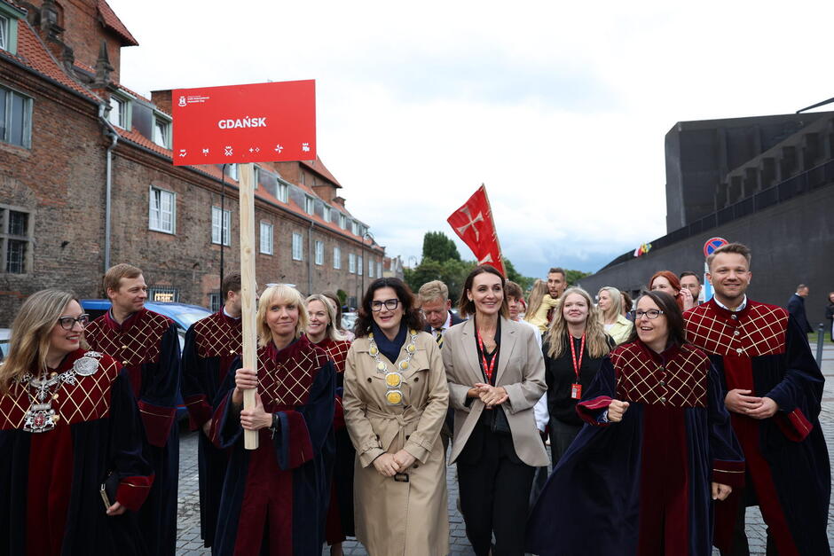 Kilkanaście osób, w tym kilka ubranych w odświętne togi, podczas parady. Jedna z nich niesie czerwoną tablicę z napisem Gdańsk