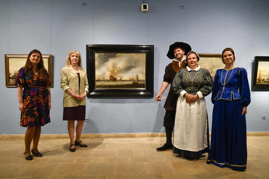 Na zdjęciu widzimy pięć osób pozujących przed obrazami w galerii sztuki. Dwie z nich są ubrane w współczesne ubrania, a trzy w stroje historyczne z epoki 