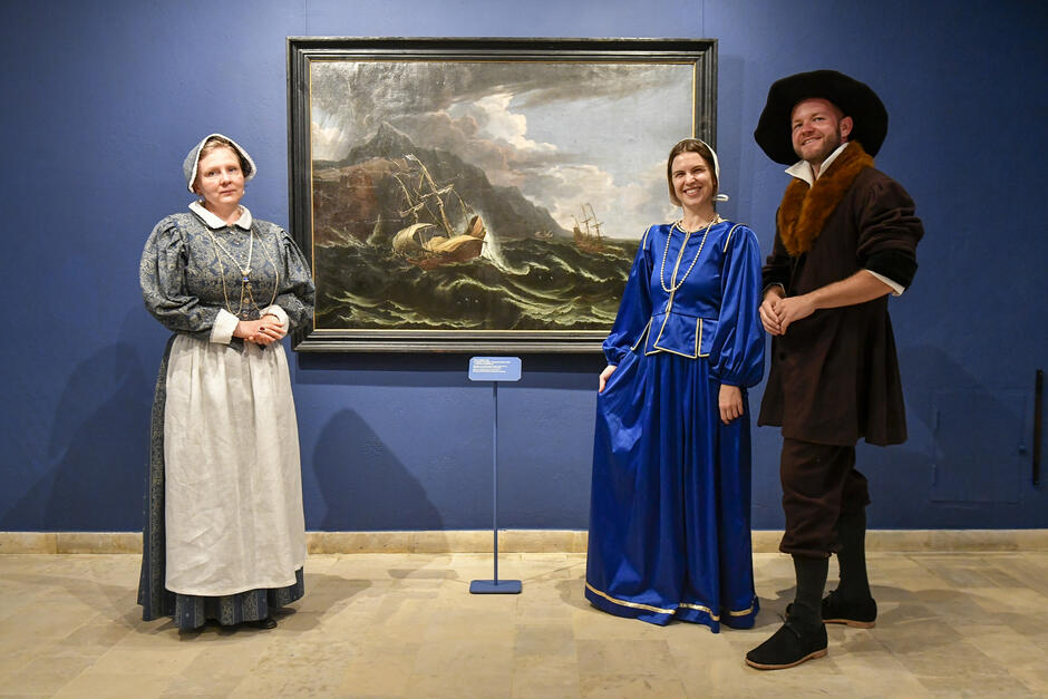 Na zdjęciu widzimy trzy osoby pozujące przed obrazem przedstawiającym scenę morską w galerii sztuki. Wszystkie osoby są ubrane w historyczne stroje z epoki