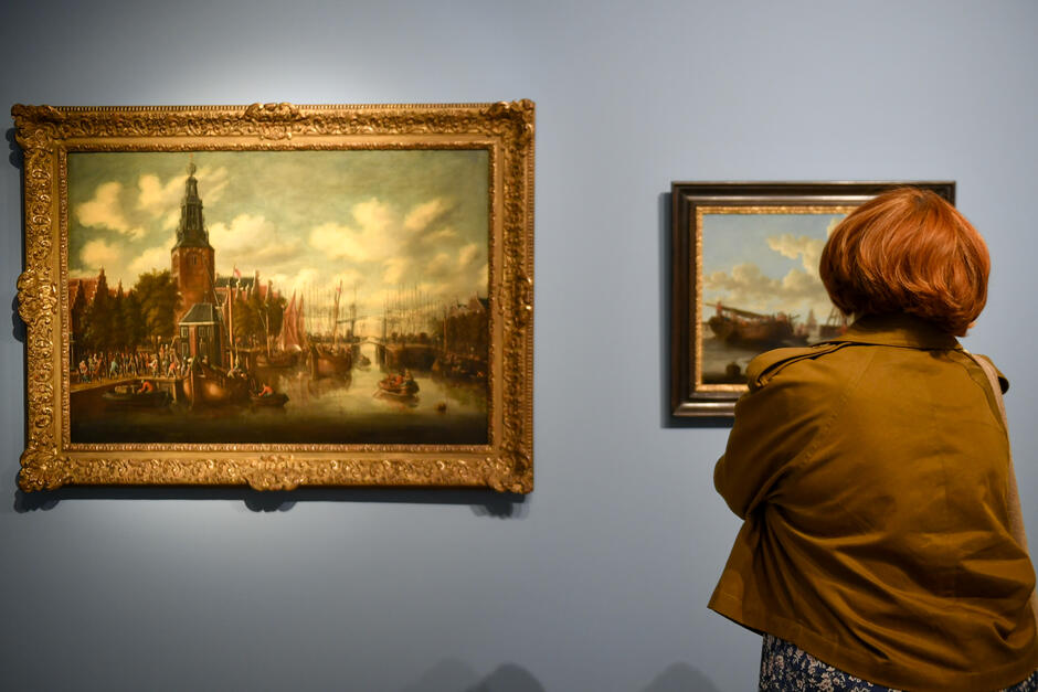 Na zdjęciu widzimy osobę z rudymi włosami, oglądającą dwa obrazy w galerii sztuki. Duży obraz po lewej stronie przedstawia scenę portową z kościołem w tle, podczas gdy mniejszy obraz po prawej stronie ukazuje statek na tle nieba z chmurami.