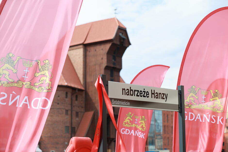Zdjęcie przedstawia uliczny znak nabrzeże Hanzy  w Gdańsku na tle charakterystycznej architektury ceglanej. Obok znaku widoczne są czerwone flagi z herbem Gdańska, na których widnieją złote lwy trzymające tarczę z krzyżem.