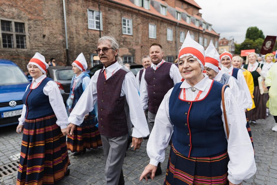 Zdjęcie przedstawia grupę ludzi w tradycyjnych strojach ludowych, uczestniczących w paradzie na bruku w historycznej części miasta. Uśmiechnięci uczestnicy, w tym starsze osoby, maszerują radośnie, trzymając się za ręce, co dodaje wydarzeniu atmosfery wspólnoty i radości.