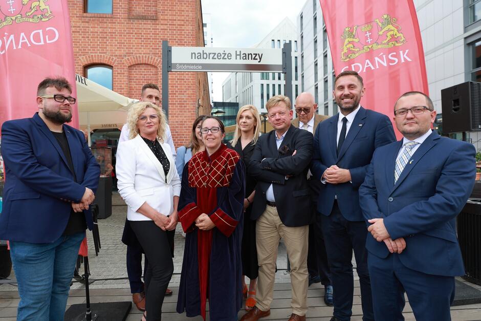 Zdjęcie przedstawia grupę oficjalnych przedstawicieli pozujących przed znakiem nabrzeże Hanzy  w Gdańsku, na tle nowoczesnych budynków i czerwonych flag miasta. Wszyscy są elegancko ubrani, a atmosfera wydaje się być uroczysta, podkreślając wagę wydarzenia.