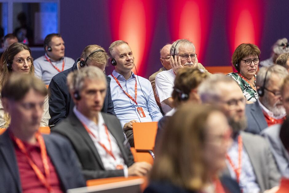 Na zdjęciu znajduje się grupa ludzi siedzących na konferencji, wszyscy mają na sobie słuchawki. Tło jest oświetlone ciepłymi, czerwonymi światłami, a większość uczestników nosi identyfikatory na czerwonych smyczach.