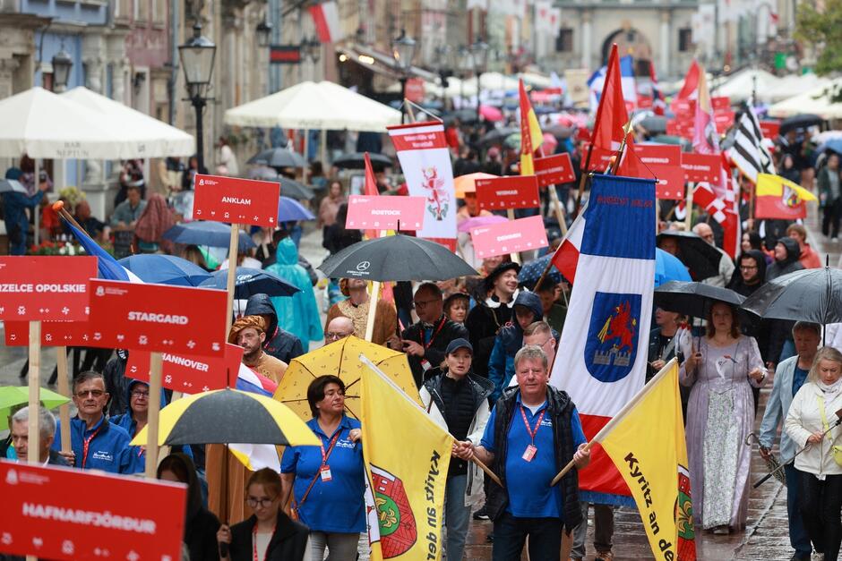 Na zdjęciu widać tłum ludzi biorących udział w marszu lub paradzie, idących ulicą w deszczu z parasolami. Wiele osób trzyma tabliczki z nazwami miast, takich jak Anklam, Stargard, Hannover i Rostock, oraz flagi, co sugeruje, że jest to międzynarodowe wydarzenie lub festiwal.