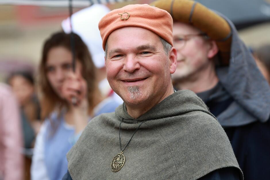 Na zdjęciu widać uśmiechniętego mężczyznę w średniowiecznym stroju, noszącego brązowy beret i medalion na szyi. W tle widać innych uczestników wydarzenia, którzy również są ubrani w historyczne kostiumy, co sugeruje, że biorą udział w rekonstrukcji historycznej lub festiwalu.