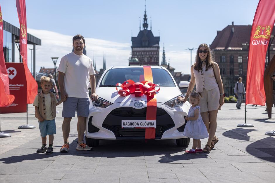 Na zdjęciu widać rodzinę składającą się z ojca, matki i dwójki małych dzieci, stojących przed białym samochodem marki Toyota, który jest ozdobiony dużą, czerwoną kokardą. Tablica rejestracyjna samochodu ma napis NAGRODA GŁÓWNA . W tle widoczny jest budynek z wieżą oraz dwie czerwone flagi z napisem  GDANSK  oraz herbem miasta Gdańska. Po lewej stronie zdjęcia znajduje się punkt informacji. Cała scena odbywa się na zewnątrz w słoneczny dzień, prawdopodobnie na jakimś placu miejskim w Gdańsku.