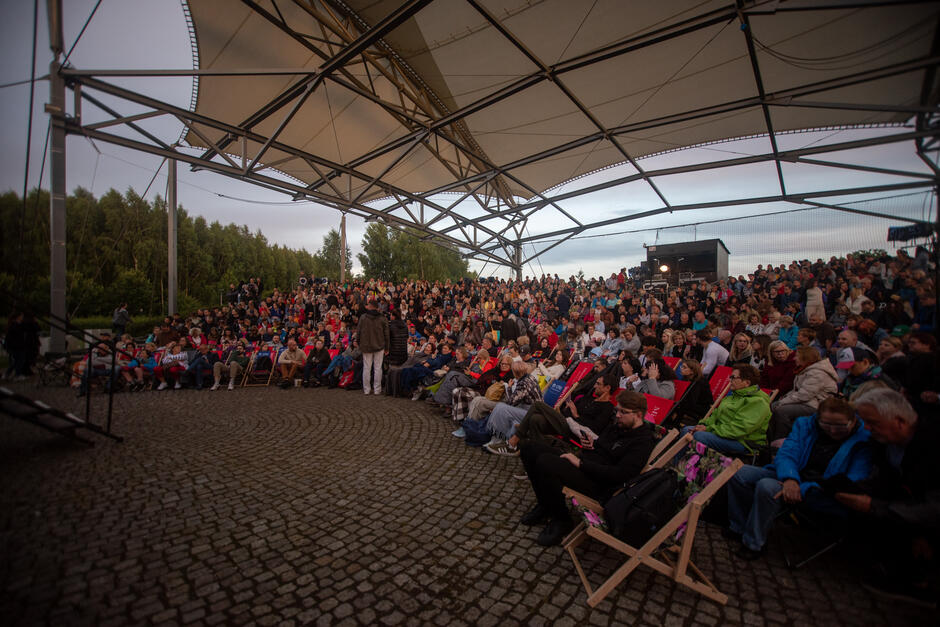 Na zdjęciu znajduje się duża publiczność zgromadzona pod zadaszoną sceną plenerową, oglądająca wydarzenie na świeżym powietrzu. Widownia składa się z wielu osób siedzących na krzesłach i leżakach, a w tle widać drzewa i wieczorne niebo, co tworzy przyjemną atmosferę