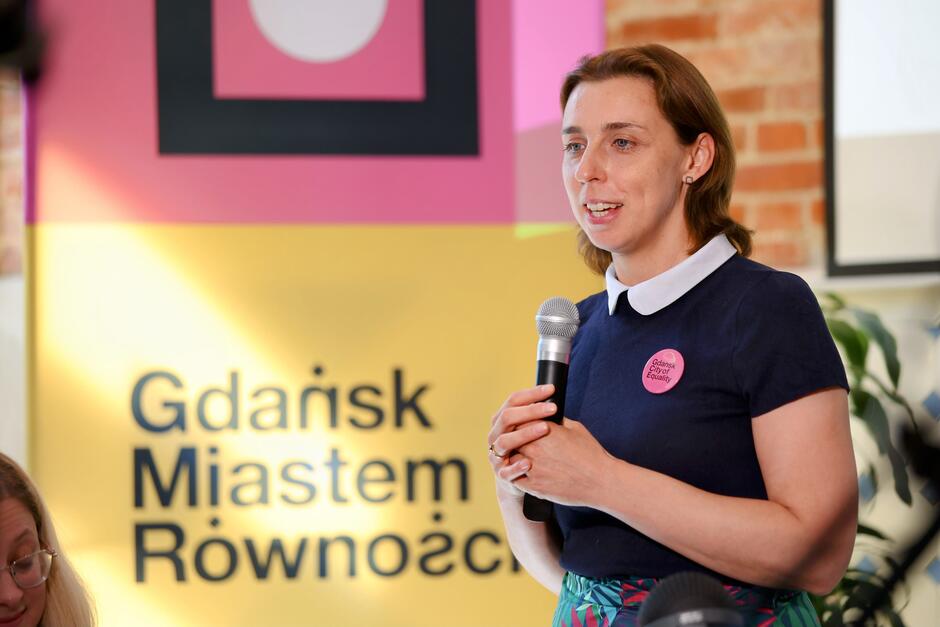 Na zdjęciu widzimy kobietę trzymającą mikrofon i przemawiającą podczas wydarzenia. W tle znajduje się kolorowy baner z napisem "Gdańsk Miastem Równości", co sugeruje, że wydarzenie jest związane z promocją równości i integracji społecznej.
