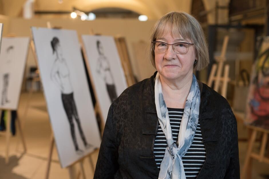 Na zdjęciu znajduje się starsza kobieta w okularach i szalu, stojąca w galerii sztuki. W tle widoczne są sztalugi z czarno-białymi rysunkami postaci