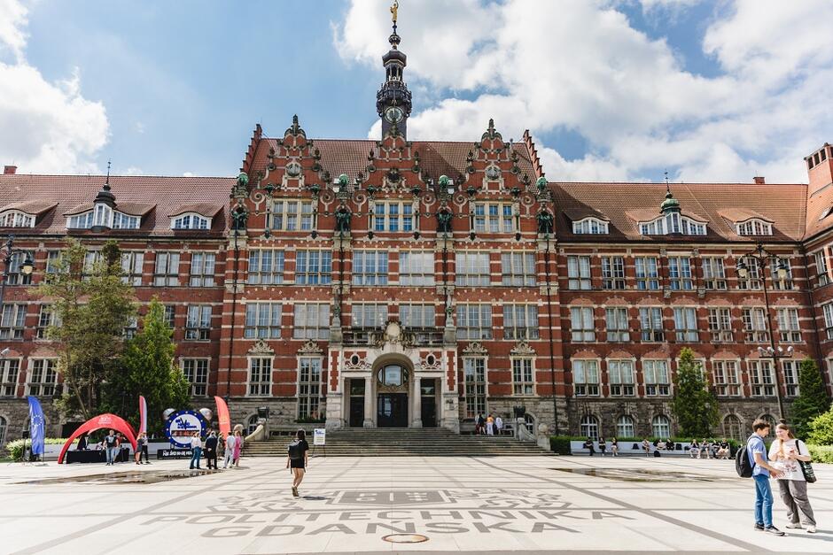 Na zdjęciu znajduje się główny budynek Politechniki Gdańskiej, charakterystyczny z czerwonej cegły i zdobionymi fasadami. Przed budynkiem widoczni są ludzie spacerujący po placu, na którym napisane jest Politechnika Gdańska 