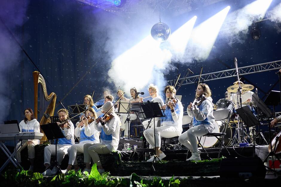 Na zdjęciu znajduje się orkiestra grająca na scenie, oświetlona intensywnym światłem reflektorów i otoczona mgłą sceniczną. Muzycy ubrani są w jasne stroje, a wśród instrumentów widać harfę, skrzypce, keyboard oraz perkusję