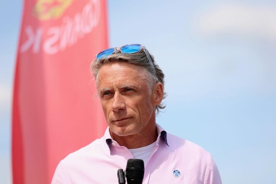 Na zdjęciu widoczny jest mężczyzna w średnim wieku z siwymi włosami, ubrany w różową koszulę z przypiętym znaczkiem i trzymający mikrofon. W tle znajduje się czerwony baner z napisem Gdańsk na tle niebieskiego nieba