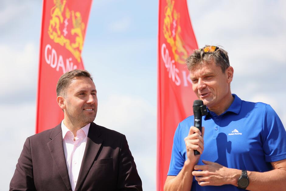 Na zdjęciu widoczni są dwaj mężczyźni, jeden w garniturze, a drugi w niebieskiej koszulce polo, trzymający mikrofon i przemawiający. W tle znajdują się dwa czerwone banery z napisem "Gdańsk" oraz herbem miasta na tle nieba.