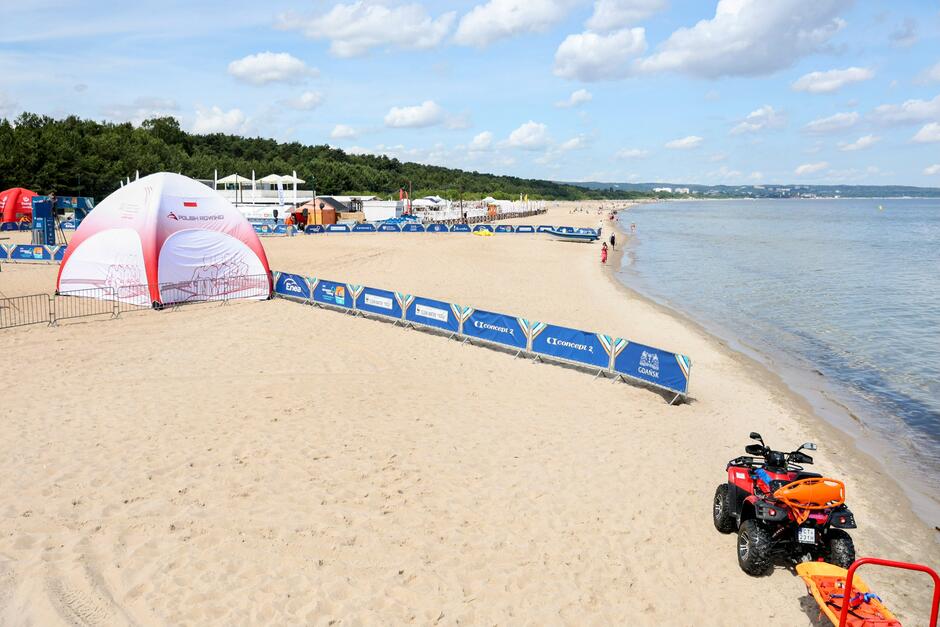 Na zdjęciu widać szeroką, piaszczystą plażę nad morzem, na której ustawiono biało-czerwony namiot z napisem "Polish Rowing" oraz liczne bariery reklamowe z logotypami sponsorów, w tym "Enea" i "Concept 2". W tle widoczne są drzewa oraz infrastruktura plażowa, a na brzegu stoi czerwoną terenówka ratownicza z pomarańczową deską ratunkową