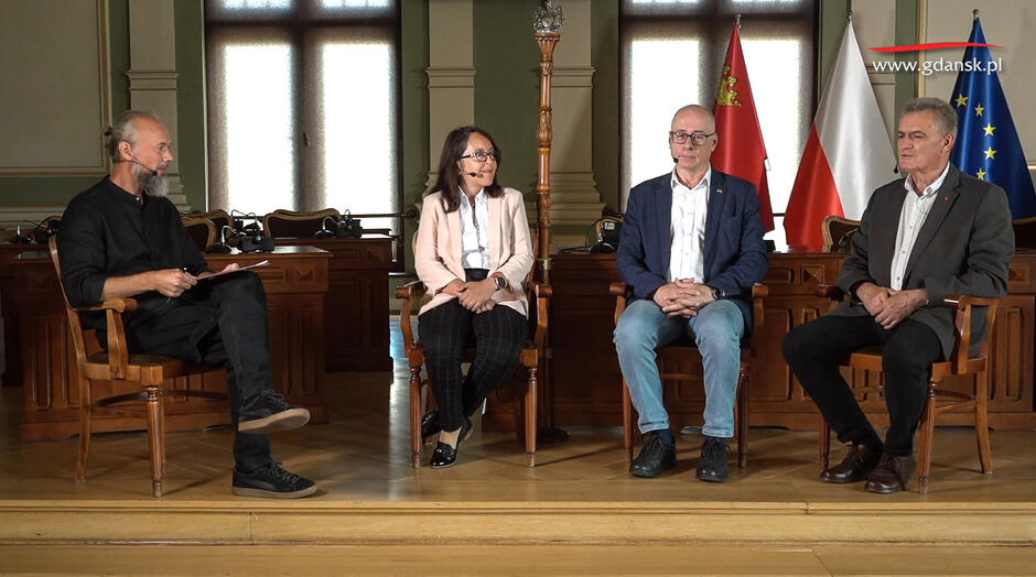 Zrzut ekranu z programu TV. Cztery osoby siedzą na podwyższeniu w dużej sali, w której obraduje Rada Miasta Gdańska. Trzech mężczyzn i kobieta mają przyczepione za uchem mikrofony i rozmawiają ze sobą.