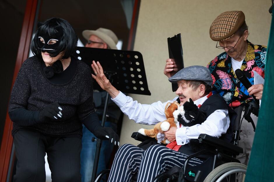 Na zdjęciu widać osoby starsze podczas występu teatralnego; jedna z nich jest przebrana za kota, a druga siedzi na wózku inwalidzkim, trzymając pluszowe kotki. Trzecia osoba w kolorowej koszuli trzyma mikrofon