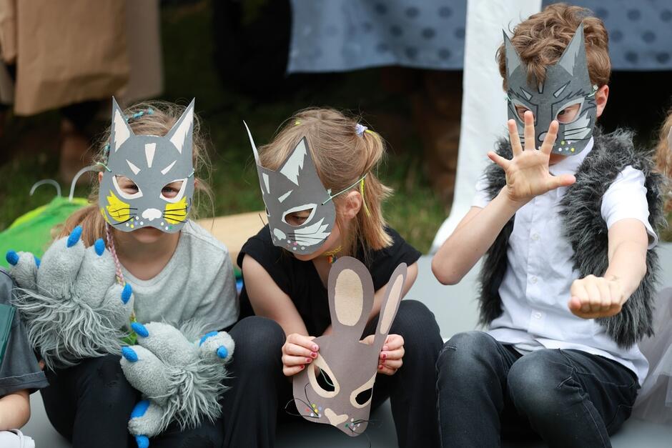 Na zdjęciu widać troje dzieci siedzących w rzędzie, ubranych w kostiumy zwierząt, z maskami kotów na twarzach. Dzieci trzymają w rękach rekwizyty, takie jak maska królika i pluszowe łapy