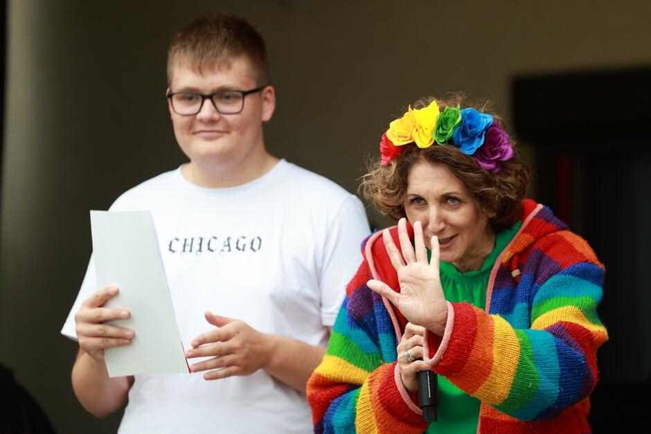 Na zdjęciu widać uśmiechniętą kobietę w kolorowym, tęczowym swetrze i wieńcu z kwiatów na głowie, trzymającą mikrofon i machającą do publiczności. Obok niej stoi młody chłopak w białej koszulce z napisem CHICAGO , trzymający kartkę papieru