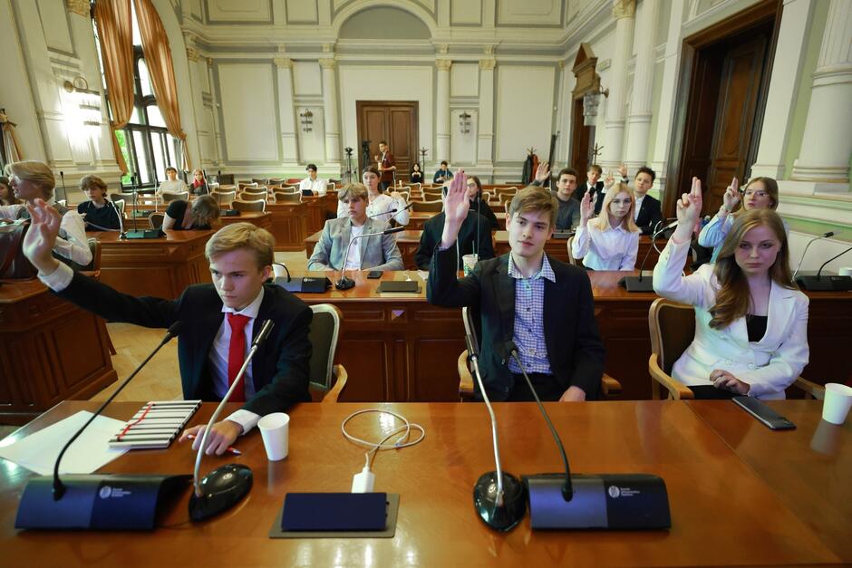 na zdjęciu kilkoro młodych ludzi, siedzą przy brązowych stołach, w kilku rzędach, podnoszą ręce do góry gdyż głosują