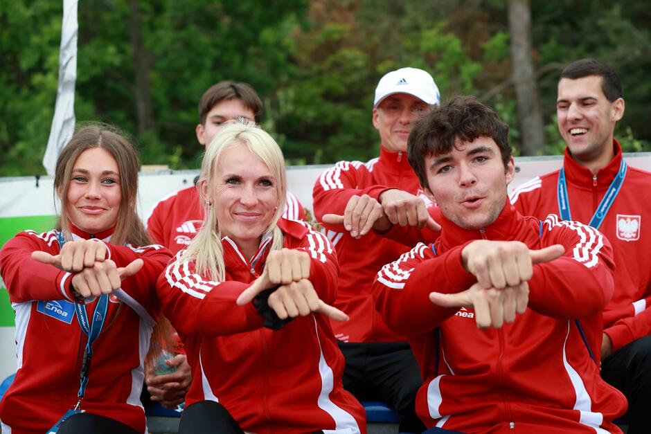 Na zdjęciu widać grupę uśmiechniętych sportowców ubranych w czerwone dresy z polskimi emblematami. Wykonują oni gest przypominający wiosłowanie, co sugeruje, że mogą to być wioślarze.
