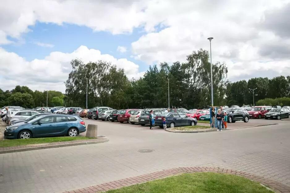 Na zdjęciu znajduje się duży parking wypełniony samochodami, położony na tle drzew i zieleni. Na pierwszym planie widać ludzi spacerujących i grupę osób rozmawiających obok zaparkowanych pojazdów
