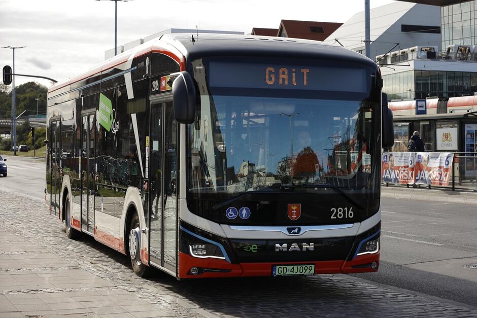 Na zdjęciu znajduje się nowoczesny autobus marki MAN z numerem rejestracyjnym GD 4J099, który należy do Gdańskiej Komunikacji Autobusowej (GAiT). Autobus stoi na przystanku, a w tle widoczne są reklamy i budynki miejskie