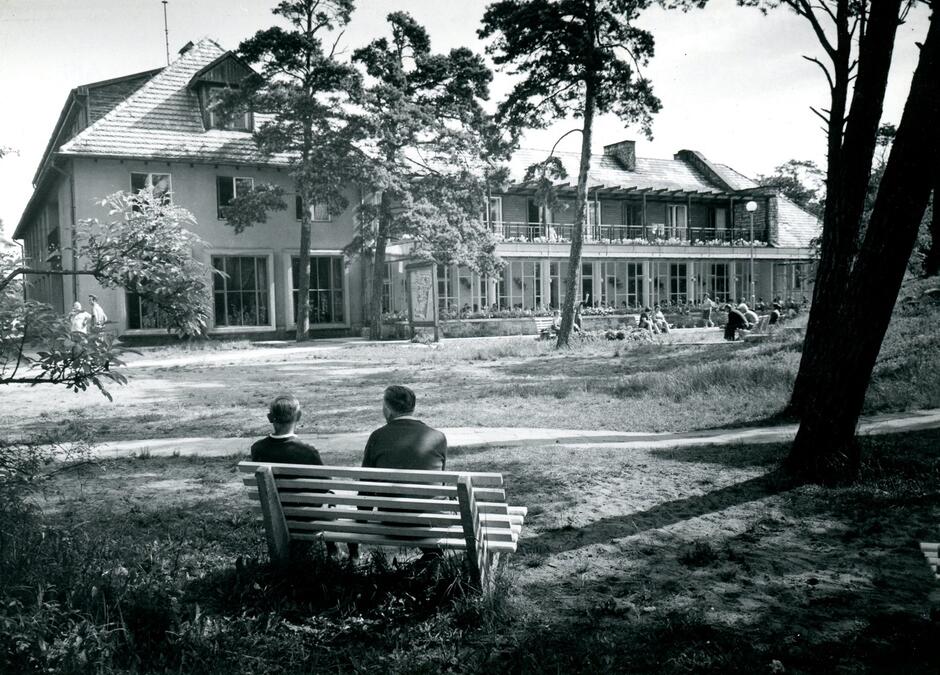 Zdjęcie przedstawia dom wypoczynkowy związku zawodowego pracowników łączności na Stogach, sfotografowany przez M. Murmana w latach 60. XX wieku. Na pierwszym planie widać dwóch mężczyzn siedzących na ławce, a w tle budynek otoczony drzewami i grupy ludzi odpoczywających na zewnątrz