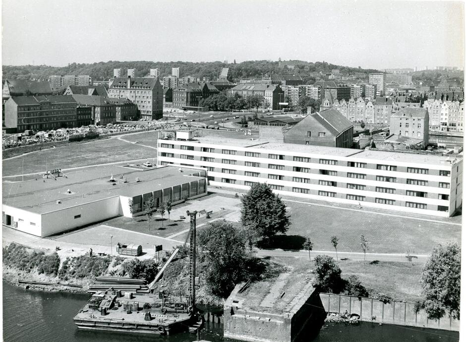 Zdjęcie przedstawia hotel Novotel sfotografowany przez K. Nowalińskiego w 1986 roku. W tle widać panoramę miasta z budynkami mieszkalnymi i przemysłowymi, a na pierwszym planie znajduje się nabrzeże z zacumowaną barką oraz teren zielony otaczający hotel