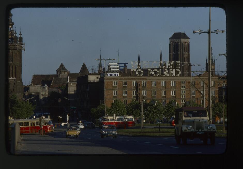 Zdjęcie przedstawia hotel Karlshof przy Kolner Chaussee 16b (obecnie ulica Spacerowa) w Oliwie na początku XX wieku. W tle widoczny jest zalesiony wzgórze z wieżą, a na pierwszym planie znajduje się spokojna ulica z kilkoma budynkami i wejściem do hotelu