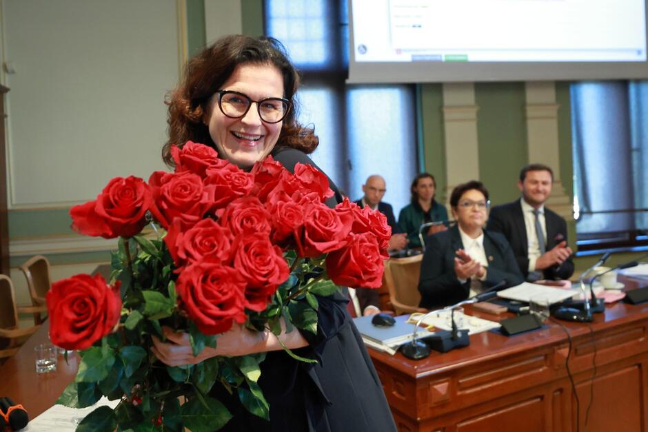 Uśmiechnięta długowłosa kobieta w okularach trzyma duży bukiet czerwonych róż 