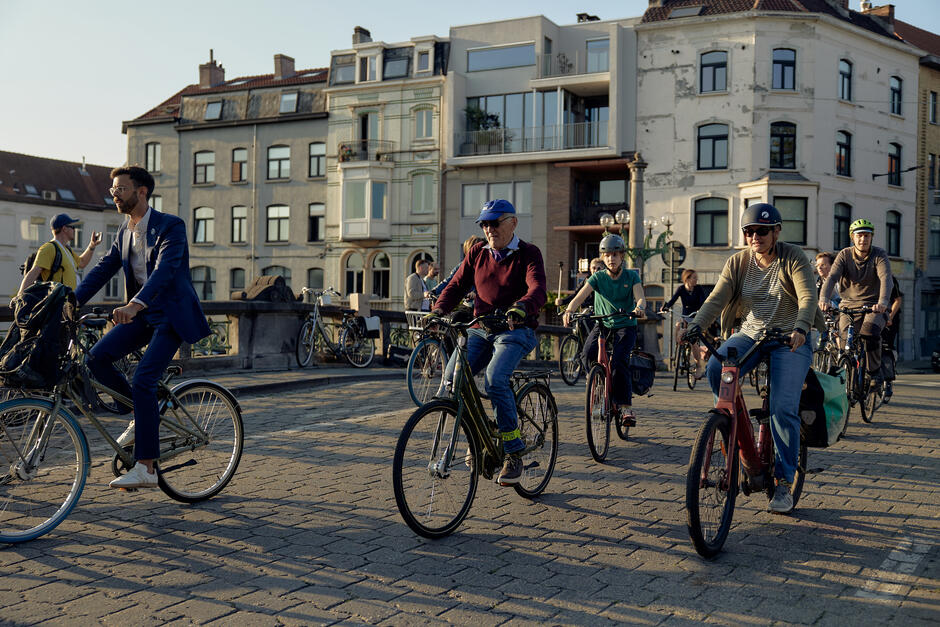 Na zdjęciu widać grupę ludzi jadących na rowerach przez brukowaną ulicę w mieście. W tle znajdują się kamienice o zróżnicowanej architekturze, a rowerzyści, wśród których są osoby w różnym wieku, są ubrani zarówno w codzienne stroje, jak i bardziej formalne ubrania.