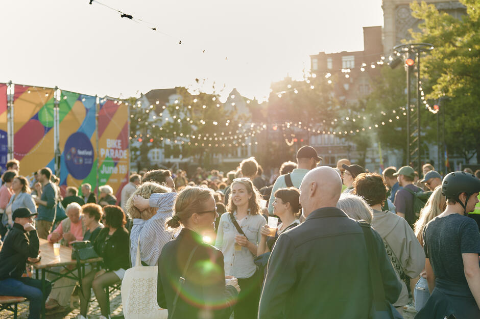 Zdjęcie przedstawia tłum ludzi zgromadzonych na zewnątrz podczas imprezy plenerowej w słoneczny dzień. W tle widać kolorowe banery reklamujące paradę rowerową oraz dekoracyjne światełka, które dodają festiwalowego klimatu wydarzeniu.