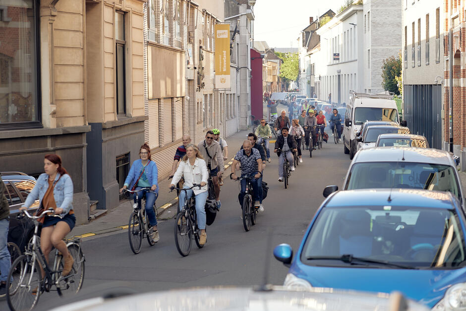 Na zdjęciu widać tłum rowerzystów jadących ulicą w mieście, otoczonych zaparkowanymi samochodami. Większość osób wygląda na zrelaksowanych, a niektórzy mają na sobie kaski ochronne.