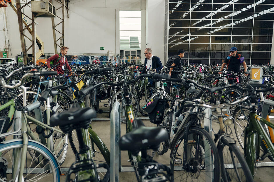 Na zdjęciu widać zatłoczony parking rowerowy w hali, gdzie wiele osób szuka swoich rowerów. W tle widać szklane ściany i dużą ilość zaparkowanych rowerów, a ludzie są zajęci odnajdywaniem swojego sprzętu wśród wielu innych.
