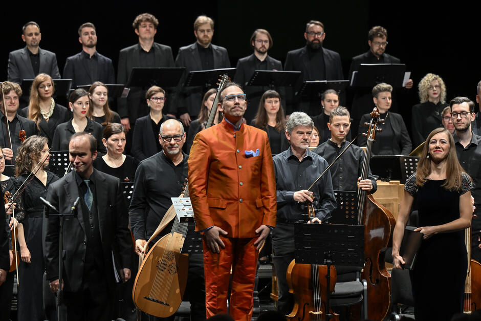 Na zdjęciu widzimy grupę muzyków, w tym dyrygenta w pomarańczowym garniturze, stojących na scenie po występie, otoczonych przez chór i orkiestrę. Wszyscy ubrani są elegancko, głównie na czarno, a ich twarze wyrażają powagę i zadowolenie