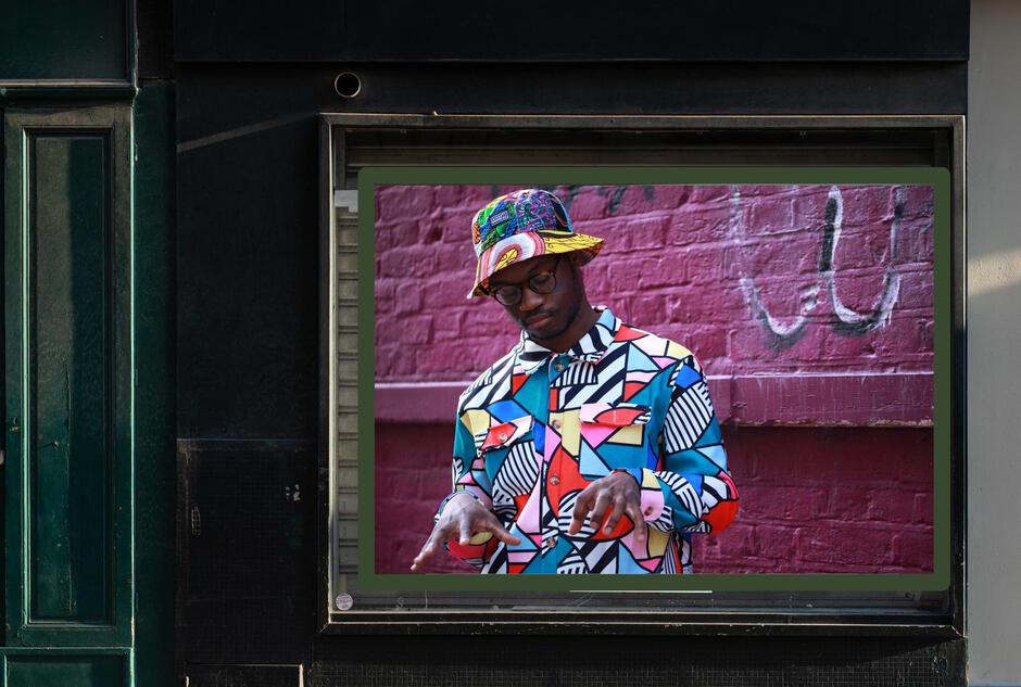 Na zdjęciu widzimy witrynę sklepową z wyświetlanym dużym zdjęciem mężczyzny w kolorowej, wzorzystej koszuli i kapeluszu. Mężczyzna pozuje na tle różowej ceglanej ściany z graffiti, mając zrelaksowany wyraz twarzy i dłonie uniesione w artystycznym geście