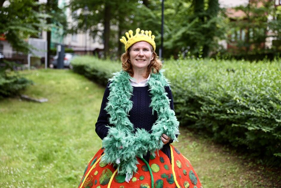 Uśmiechnięta kobieta w nakryciu głowy w kształcie złotej korony stoi na tle alei zieleni. Ma na sobie czarny dopasowany tiszert i kolorową spódnicę.