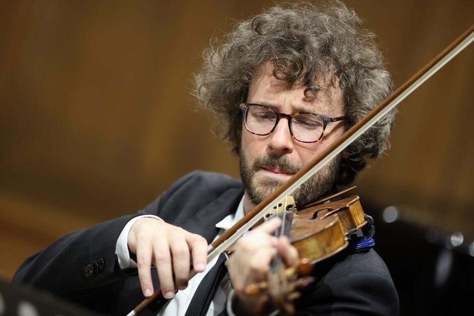 Na zdjęciu widać mężczyznę z bujnymi, kręconymi włosami i okularami, który gra na skrzypcach. Jego twarz jest skupiona