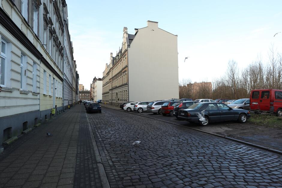 Brukowana ulica, po lewej chodnik i ciąg kamienic, po prawej parking z samochodami, za nimi budynki mieszkalne