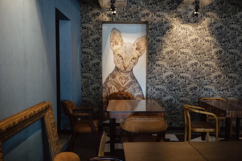 Na zdjęciu znajduje się wnętrze stylowej kawiarni lub restauracji z dużym portretem kota na ścianie w tle. W pomieszczeniu znajdują się eleganckie meble, w tym drewniane stoły i skórzane krzesła, a ściany pokryte są dekoracyjną tapetą z roślinnym wzorem
