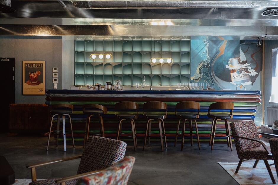 Na zdjęciu widoczny jest stylowy bar z kolorowym kontuarem i wysokimi krzesłami ustawionymi przed nim. W tle znajdują się półki o unikalnym, geometrycznym designie, a na ścianie wisi plakat reklamujący koktajl Negroni oraz mural promujący whisky Singleton