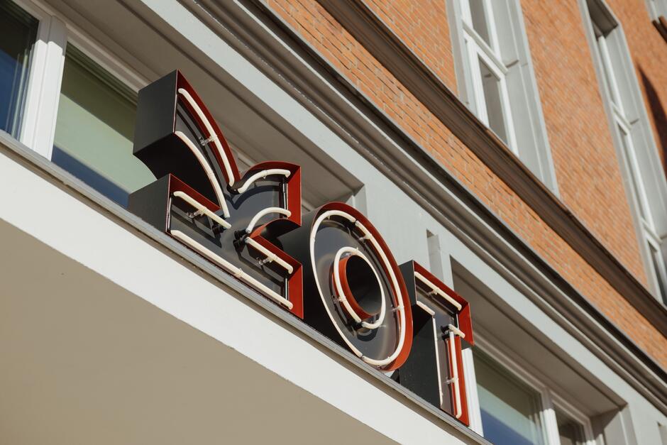 Na zdjęciu widoczny jest neonowy szyld z napisem KOT  zamontowany na elewacji budynku z cegły. Szyld jest stylizowany, a litery są w kolorach czerni i czerwieni z neonowym podświetleniem, co nadaje mu nowoczesny wygląd.
